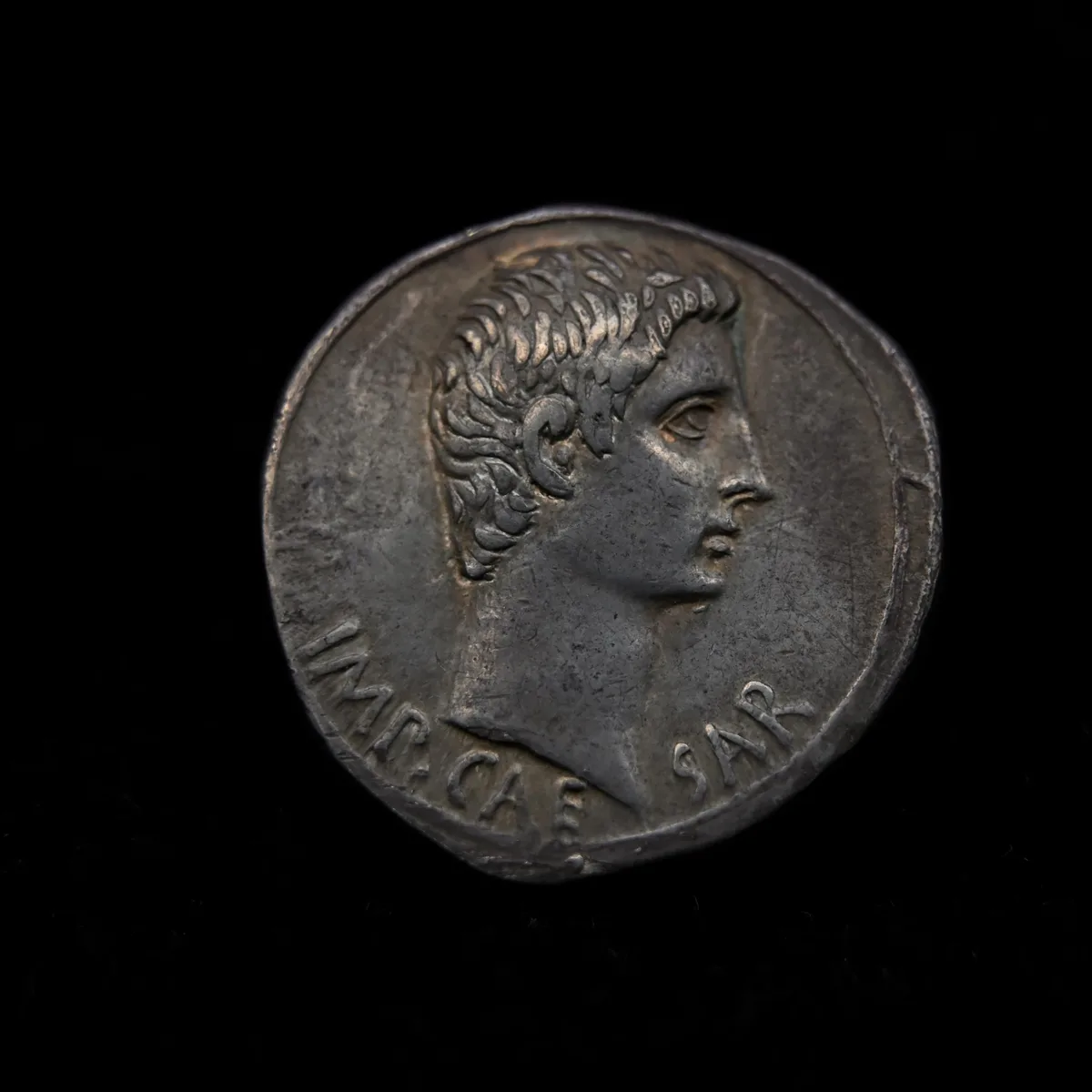 A close-up of an ancient Roman silver coin showing a profile of a man with curly hair and a laurel wreath, with the Latin…