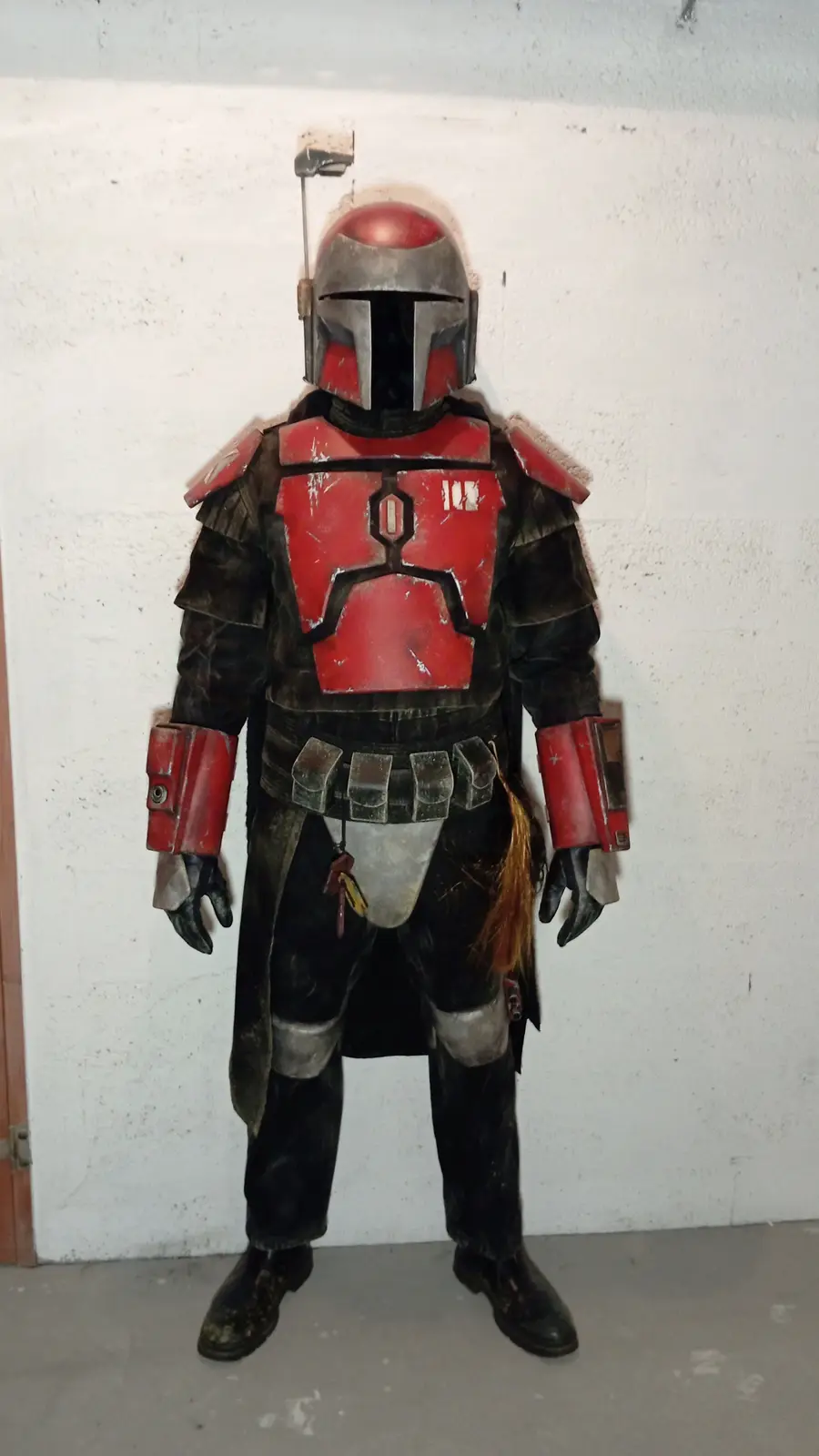 A person in a full-body Mandalorian armor costume, featuring red and black plating with a helmet, stands against a plain…