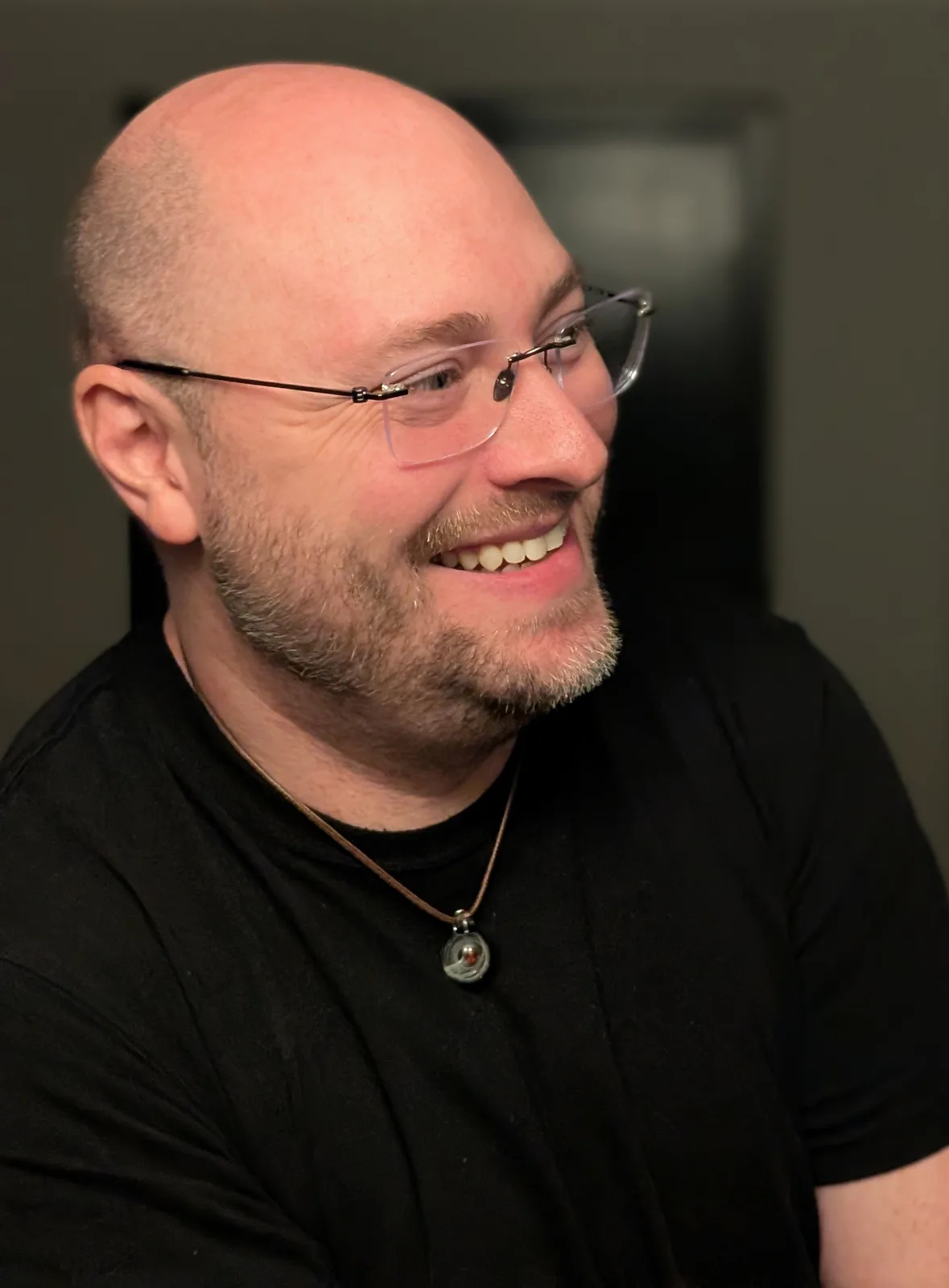 A bald man with glasses and a goatee smiles while looking off-camera, wearing a black t-shirt and a necklace with a small…