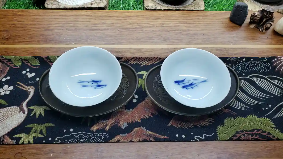 Two identical white ceramic bowls with blue fish painted inside sit on dark metal saucers atop a traditional Japanese-style…