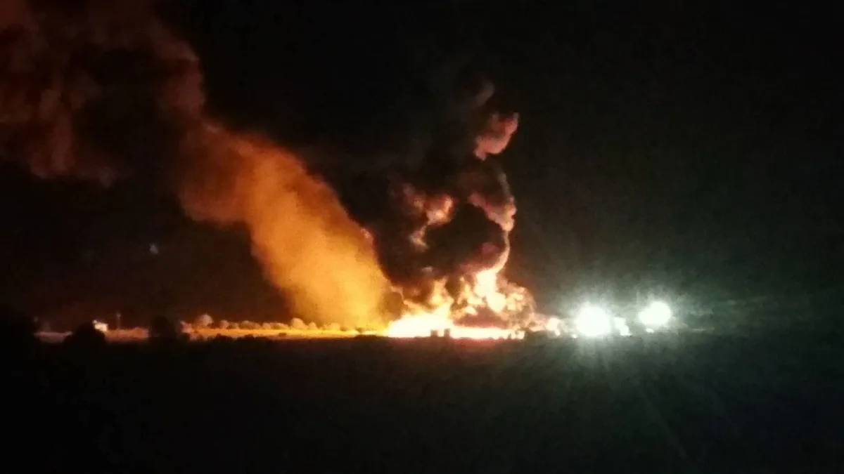 A nighttime photo shows a large fire with bright orange flames and thick smoke billowing into the dark sky, with two bright…