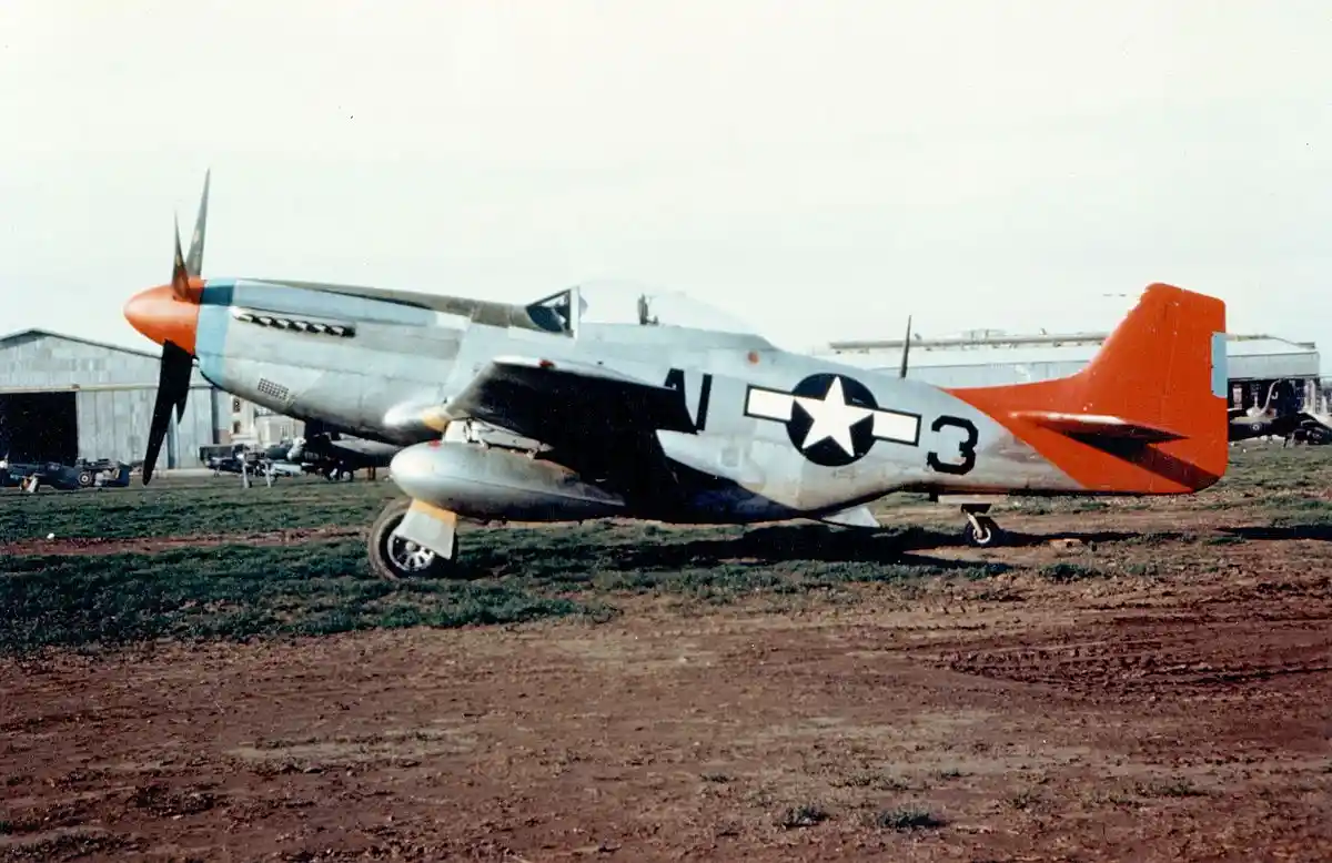 A vintage silver and orange P-51 Mustang fighter plane with U.S. military markings sits on a grassy airfield. The overlaid…