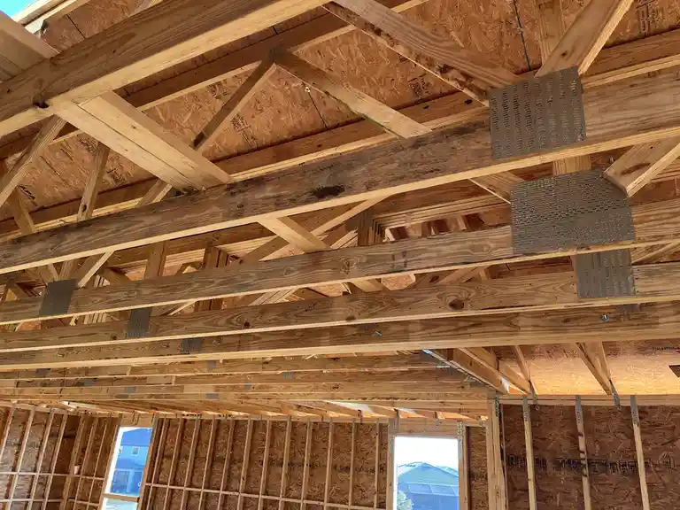 An upward-looking photo of a house under construction, showing exposed wooden beams, joists, and oriented strand board (OSB)…