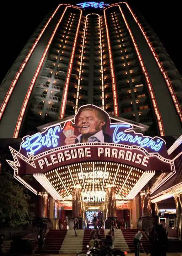 A nighttime photo of a tall, brightly lit casino building with a large, glowing sign that reads 'Buff Tannen's Pleasure…