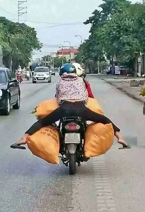 A person riding a motorcycle with two large orange sacks strapped to either side of the rear, obscuring the rider's legs…