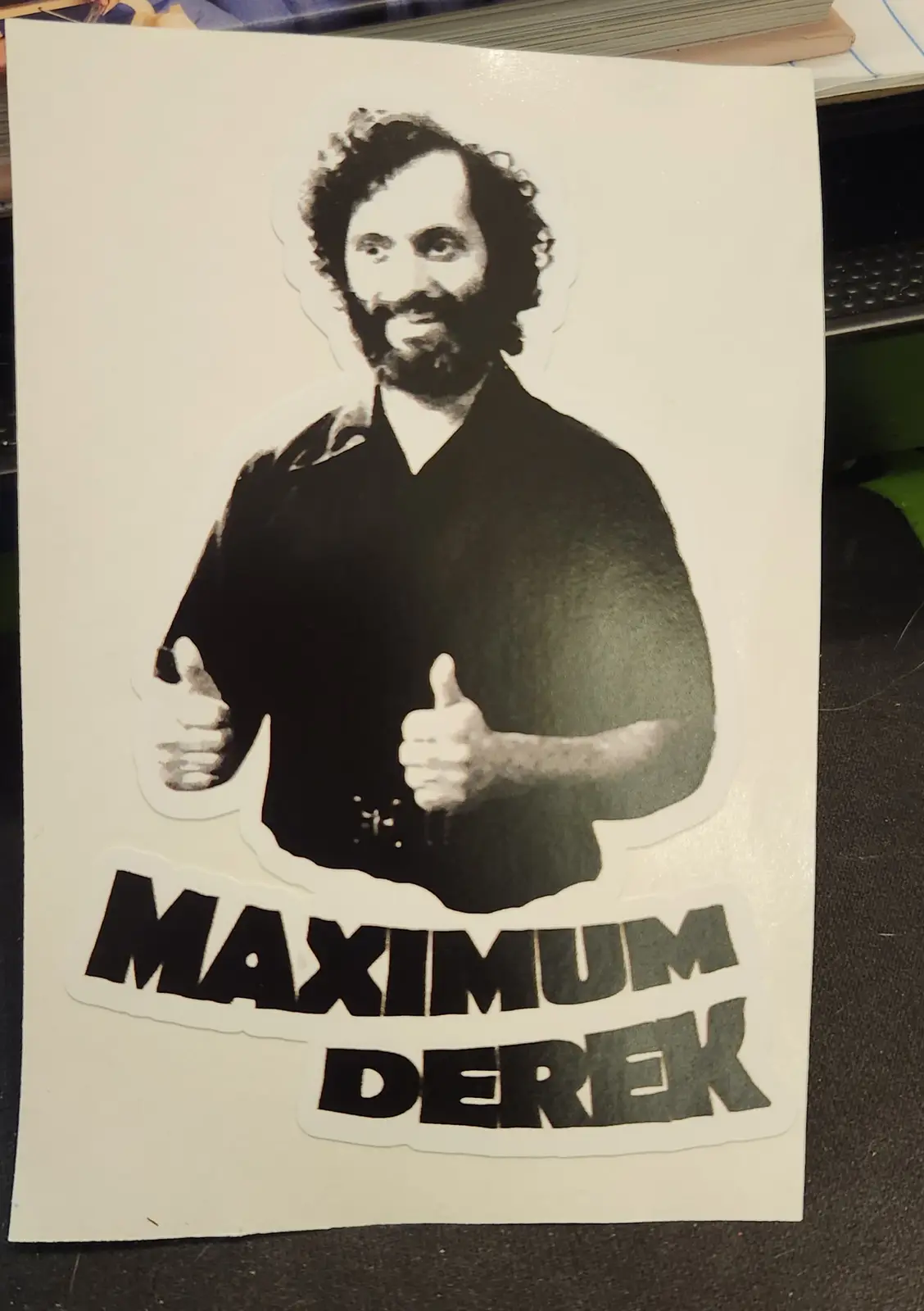A black and white sticker shows a man with curly hair and a beard giving two thumbs up. Below the image, bold text reads…