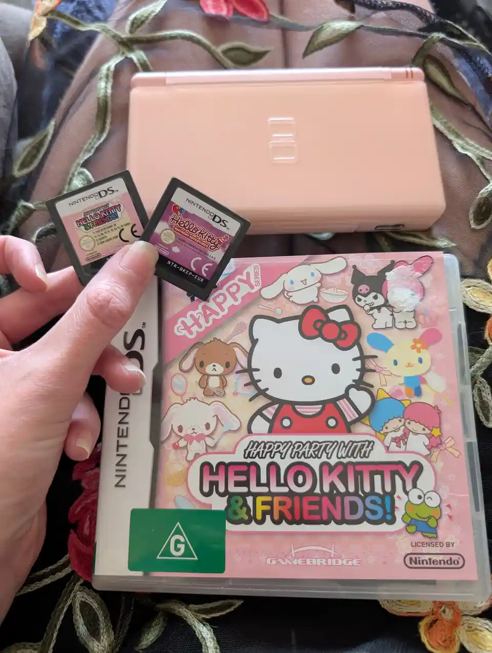A hand holds two Nintendo DS game cartridges next to the game case for 'Happy Party with Hello Kitty & Friends!' and a pink…