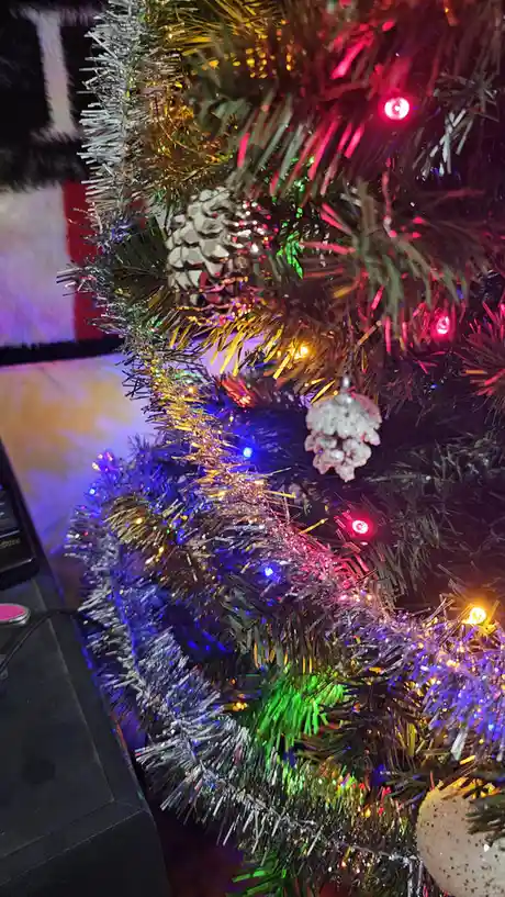 A close-up of a decorated artificial Christmas tree with colorful lights, tinsel, and pinecone ornaments. The overlaid text…