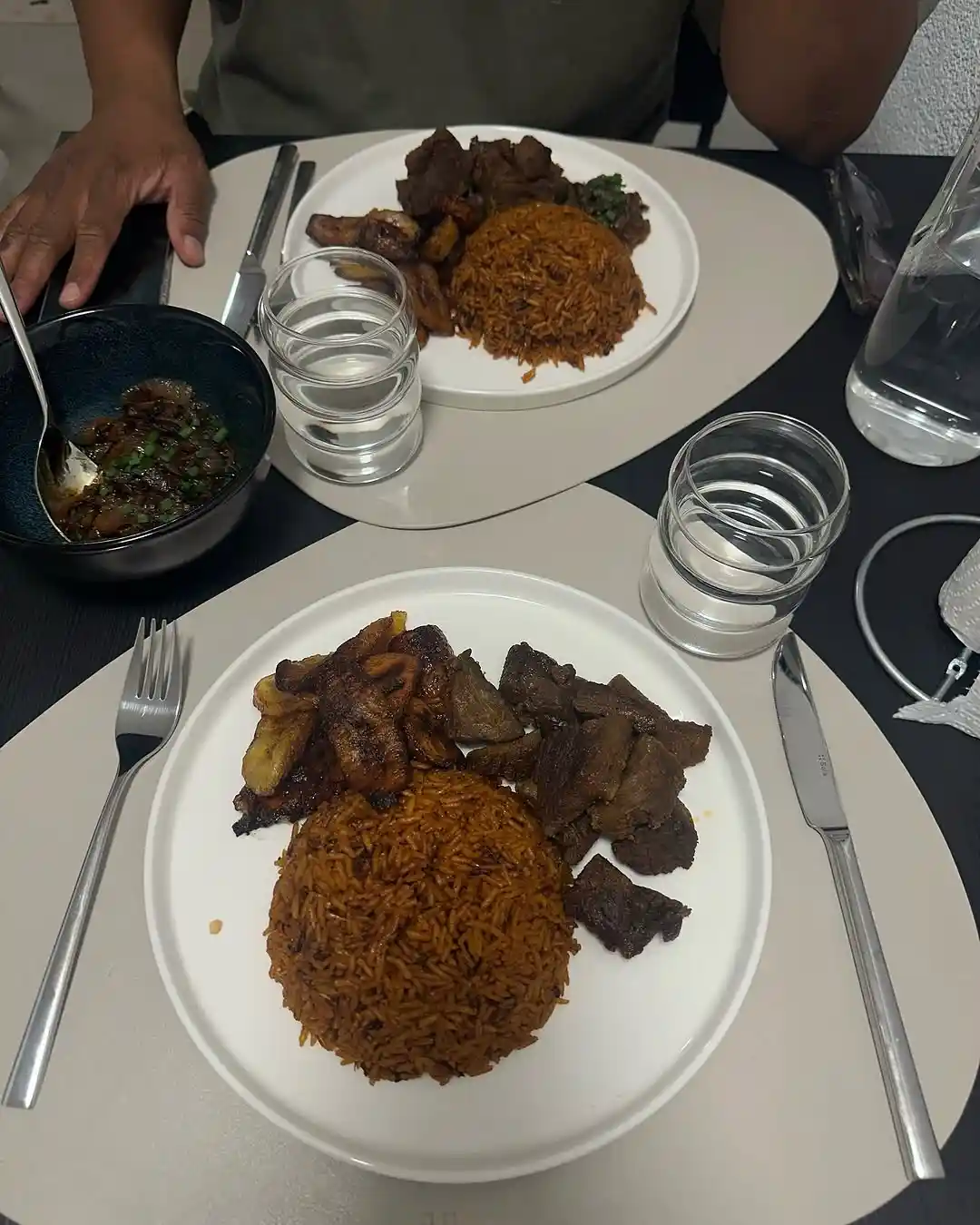 A top-down photo of two identical plates of jollof rice with meat and fried plantains, set on a dining table with glasses of…
