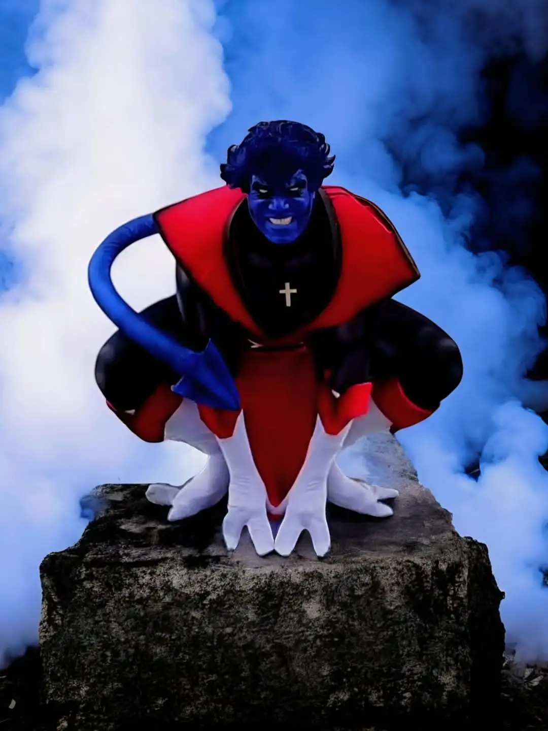 A person in a blue-skinned, red-and-black costume with a cross on their chest crouches on a rock surrounded by blue and…