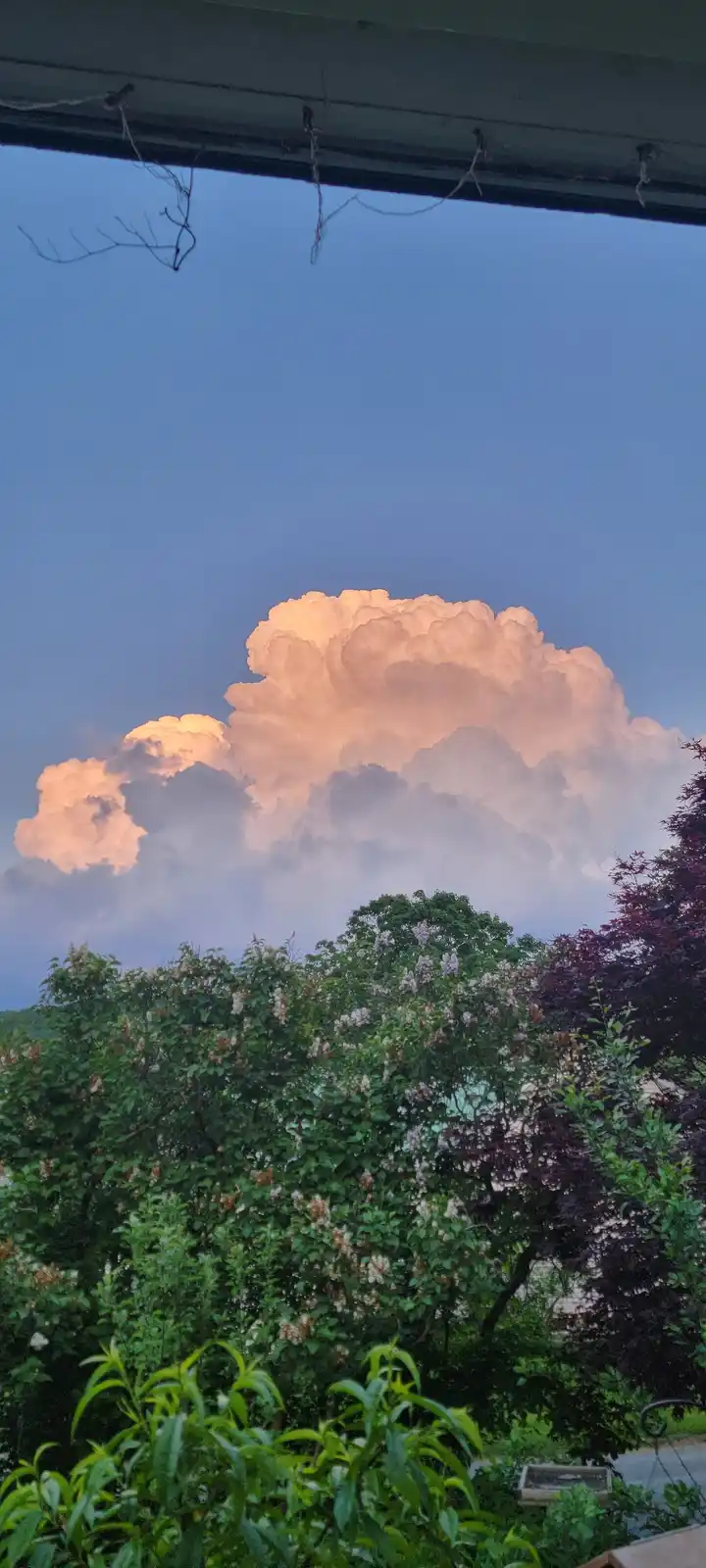 A photo taken from under a porch roof shows a large, dramatic, pink-tinged cumulonimbus cloud against a twilight sky, with…