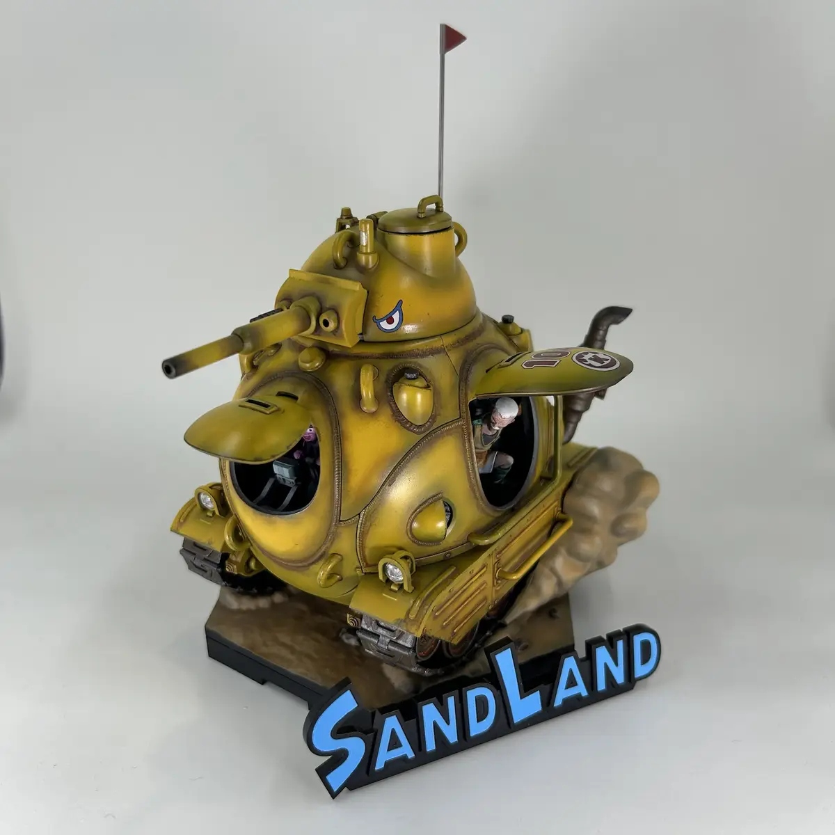 A yellow, cartoonish tank model with a single visible eye, a small red flag on top, and two miniature figures inside its…