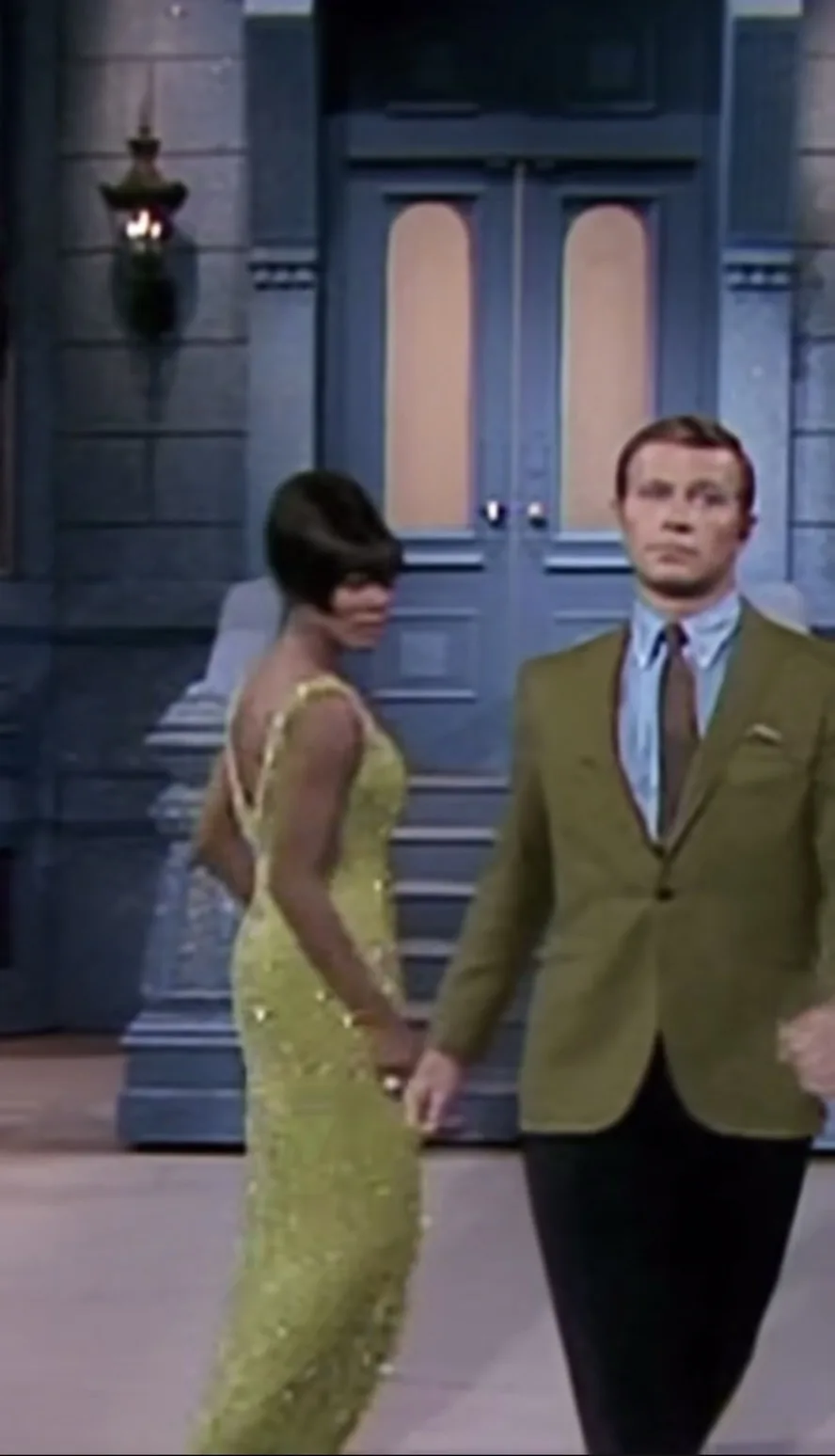 A man in a green suit and tie walks beside a woman in a sparkling yellow dress, both moving forward in a hallway with blue…