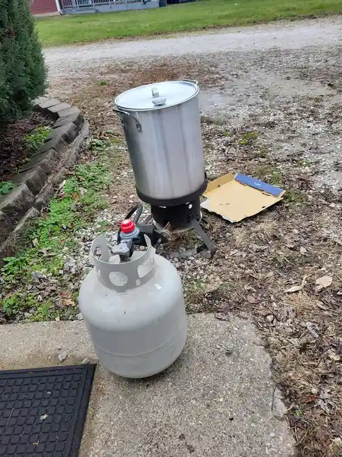 A large stainless steel pot sits atop a portable gas stove outdoors, connected to a propane tank on a concrete step. Steam…