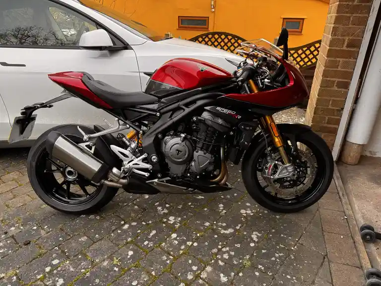 A red and black Triumph Speed Triple 800 RR motorcycle is parked on a mossy brick driveway next to a white car, with an…