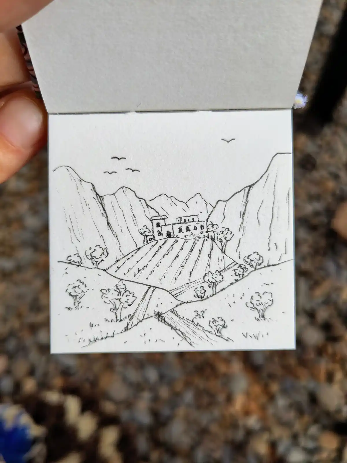 A hand holds open a small sketchbook displaying a black-and-white line drawing of a mountainous landscape with a building on…