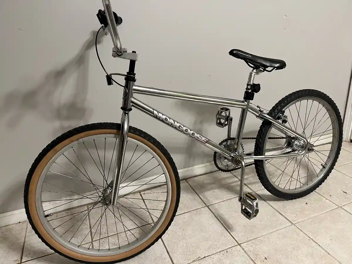A chrome-finished Mongoose BMX bike with tan-wall tires is leaning against a plain wall on a tiled floor. The brand name…
