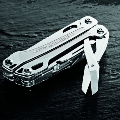 A silver Leatherman multi-tool is shown open on a dark, textured surface, with its scissors blade extended. The brand name…