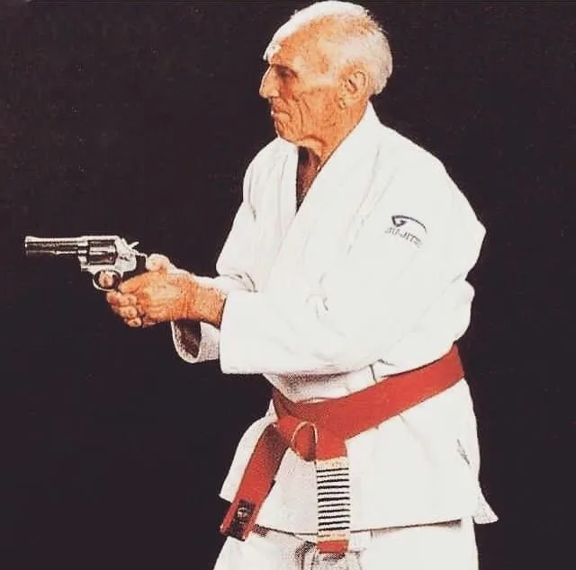 An elderly man in a white martial arts gi with a red belt holds a revolver in a ready stance against a black background. The…