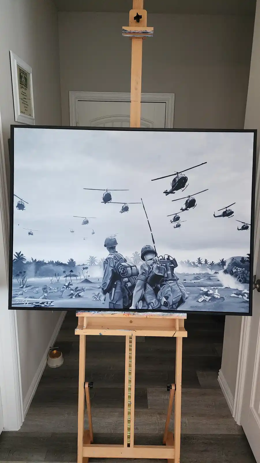 A large painting depicting soldiers and helicopters in a war scene is displayed on a wooden easel in a hallway. The painting…