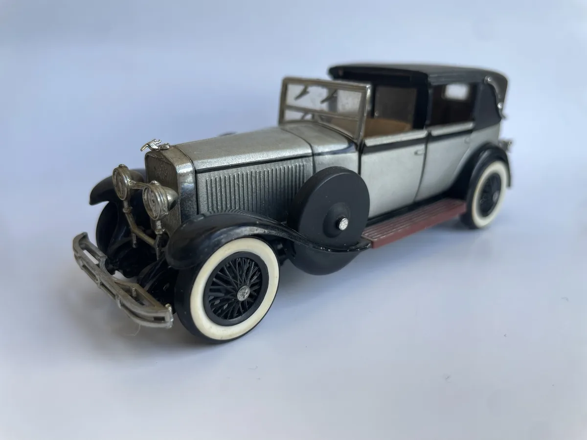 A silver and black vintage toy car model with white-walled tires and a spare tire mounted on the side, photographed against…