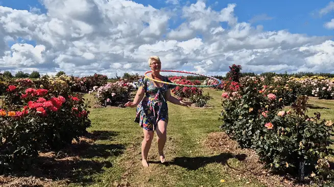 A woman in a patterned dress is shown in a rose garden under a cloudy sky, hula hooping in three sequential frames on the…