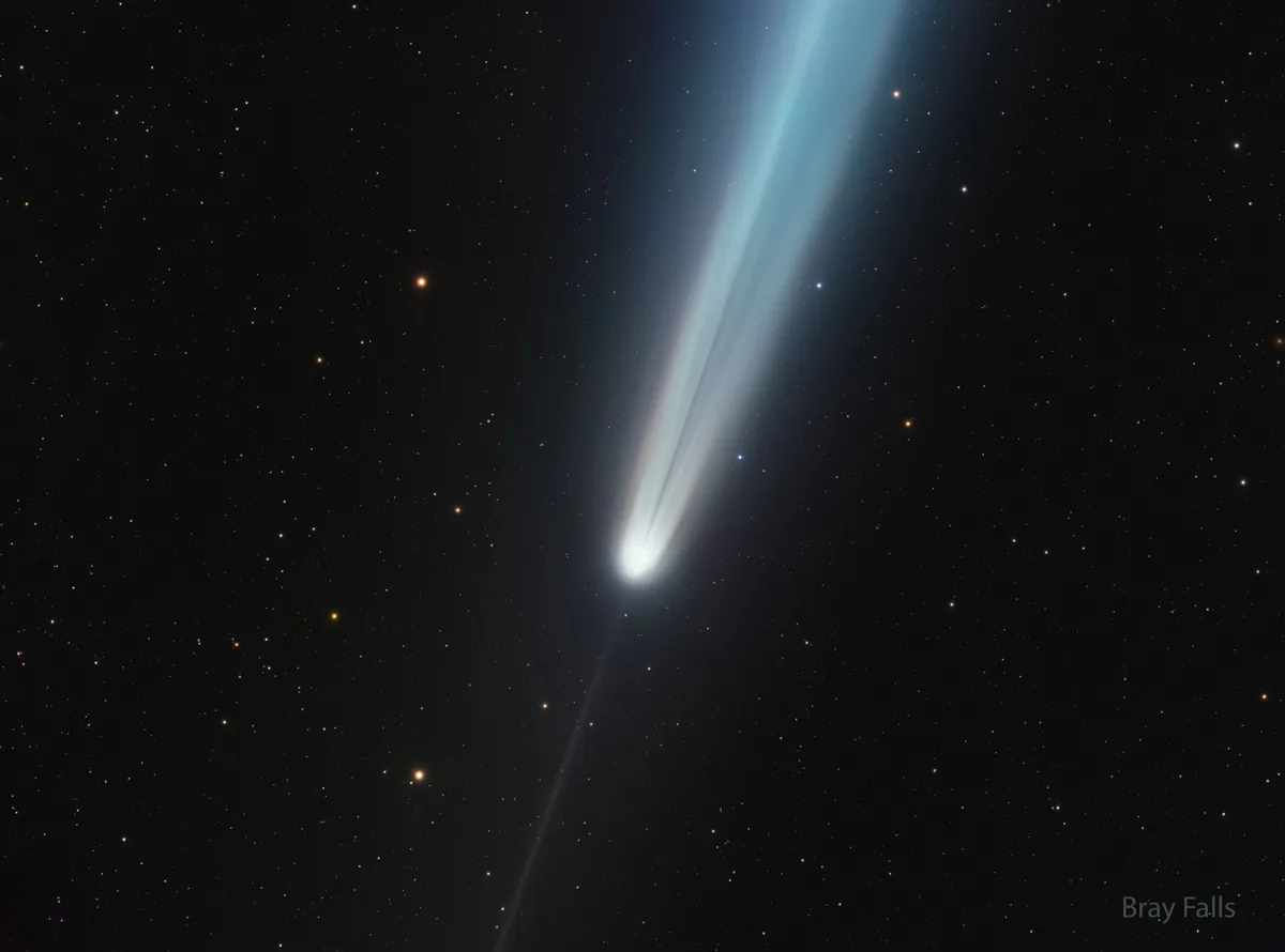 A celestial photograph shows a bright comet with a long, glowing blue and white tail streaking across a star-filled night…