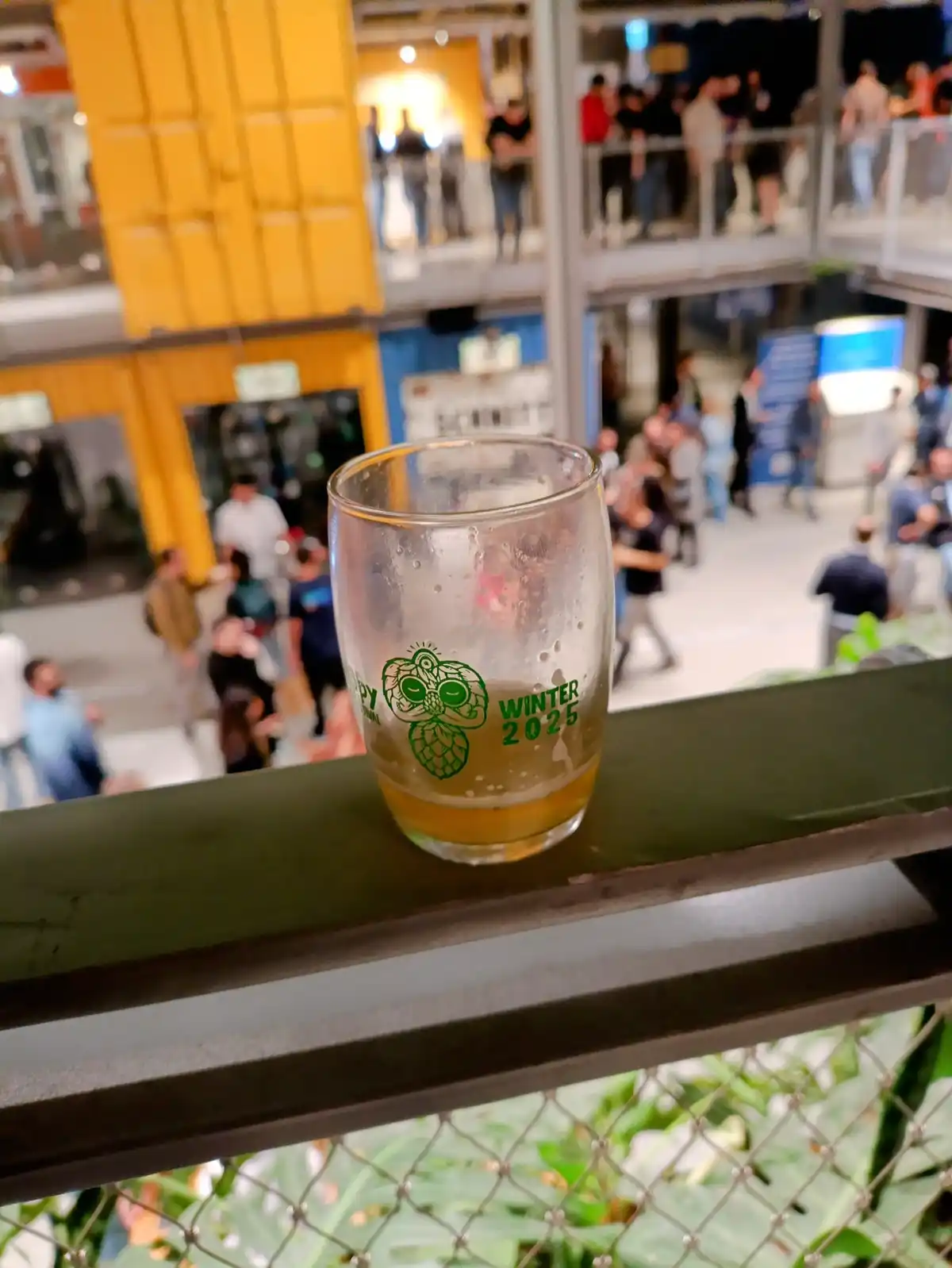 A nearly empty beer glass with a green hop logo and the text 'WINTER 2025' sits on a railing overlooking a crowded…