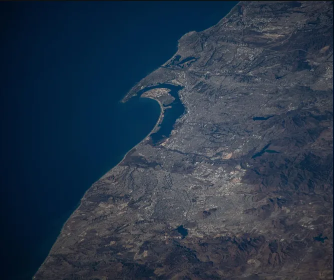 An aerial satellite image shows a coastal city nestled between deep blue ocean and rugged brown mountains, with visible…