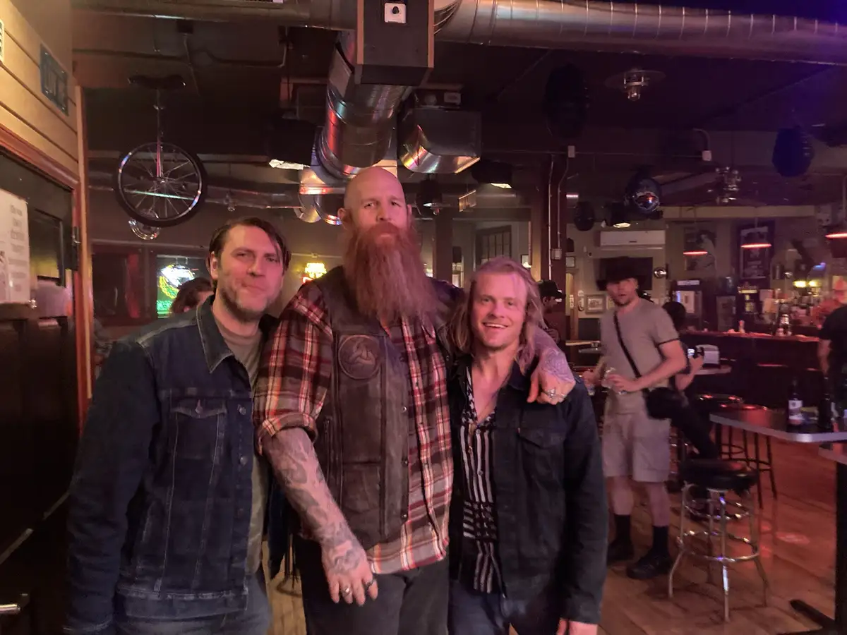 Three men pose for a photo in a dimly lit bar with exposed ductwork and a unicycle hanging from the ceiling. The man in the…