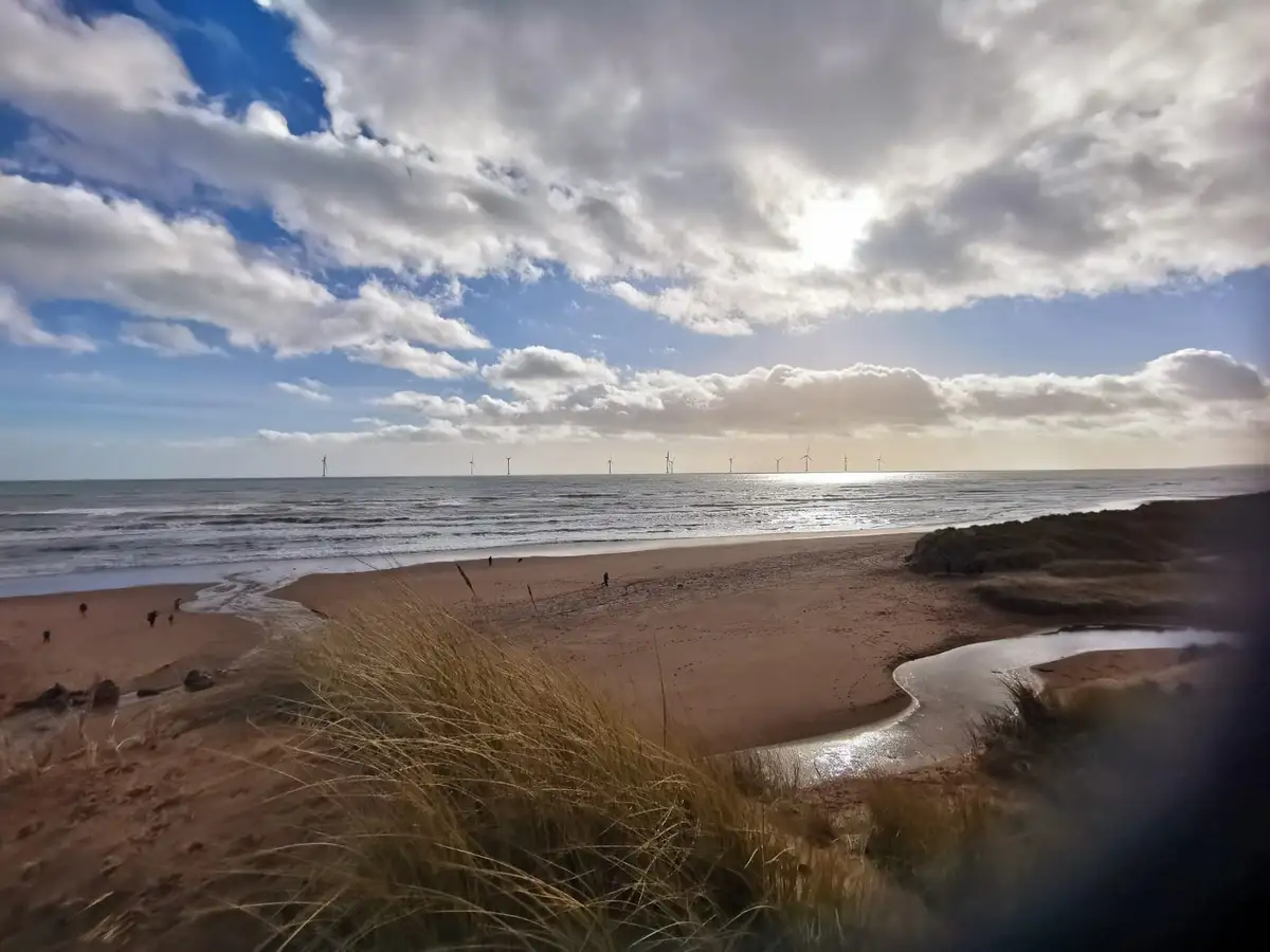 A scenic beach photo with wind turbines visible on the horizon over the ocean, under a partly cloudy sky. Several people are…