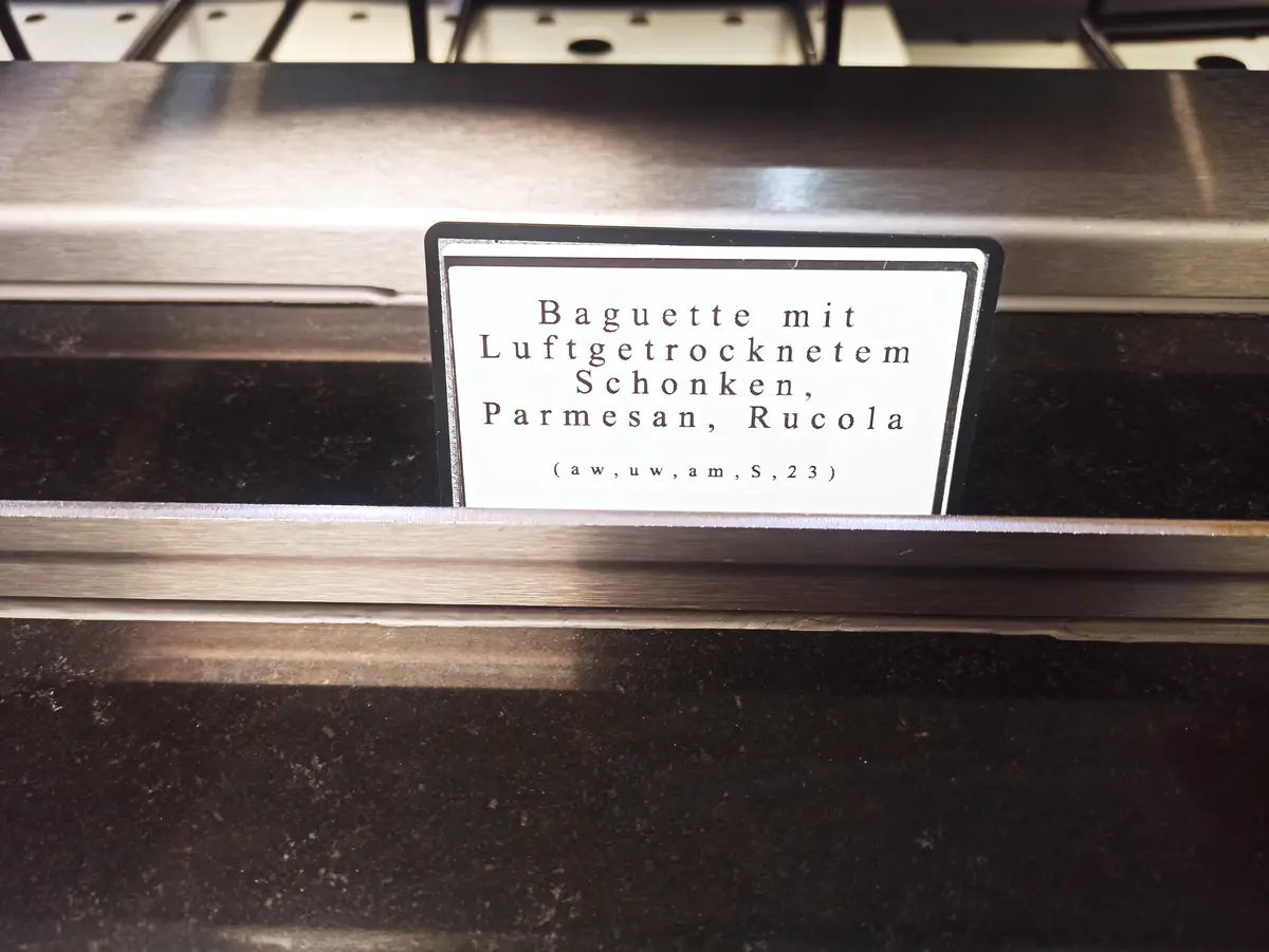 A close-up of a white label with black text on a dark shelf inside a display case, likely in a deli or bakery. The label…