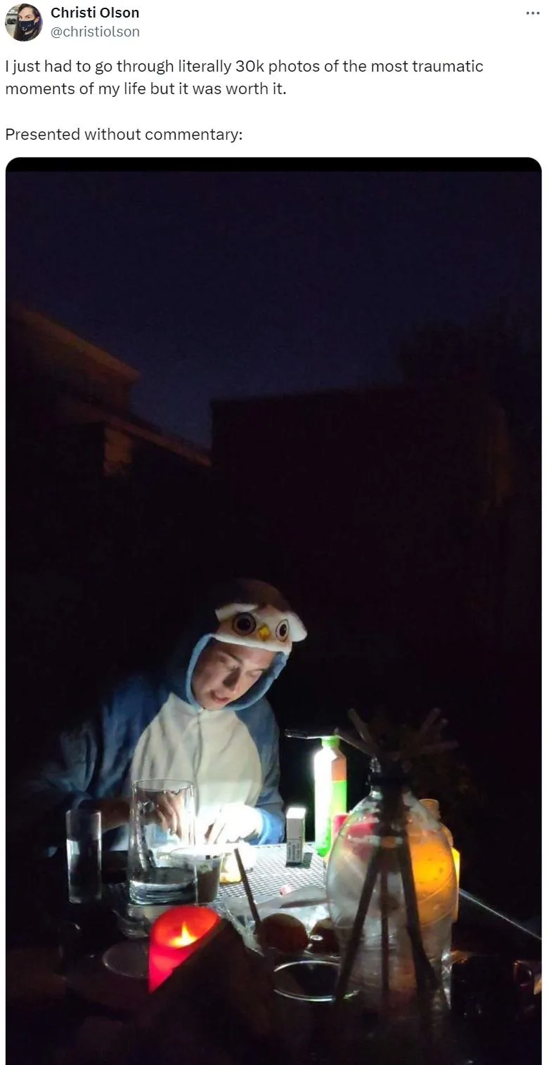 A person wearing an owl-themed hooded onesie sits at a dimly lit outdoor table at night, illuminated by a small lamp and a…