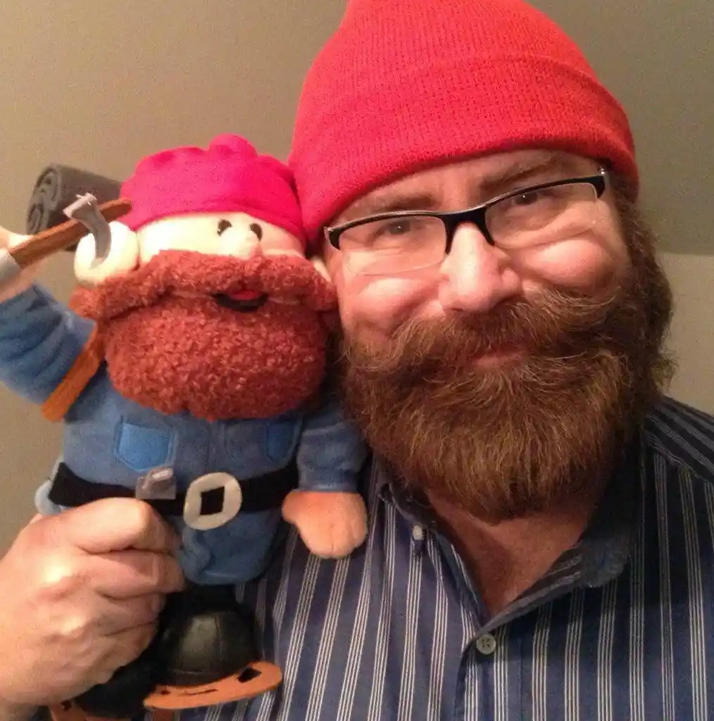 A man with a full beard, glasses, and a red beanie holds a plush toy resembling himself, which also has a red beard…