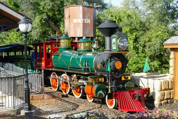 A vintage green and red steam locomotive with orange wheels is parked on tracks next to a large rusty water tank labeled…