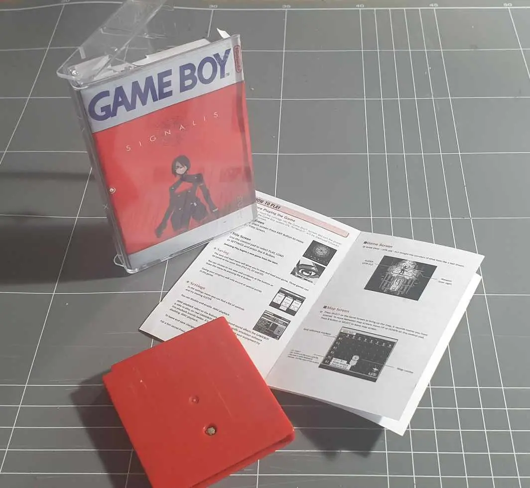 A red Game Boy cartridge labeled 'SIGNALIS' sits in a clear plastic case next to its open instruction manual and a red…