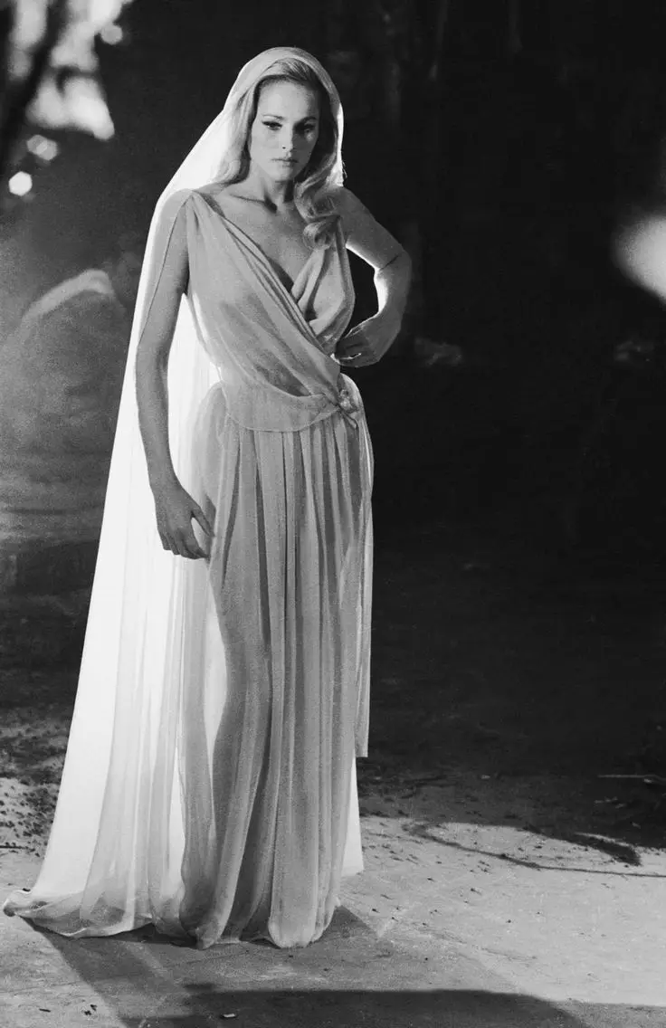 A black-and-white photo of a woman in a flowing, draped wedding gown with a veil, standing outdoors at night with a serious…