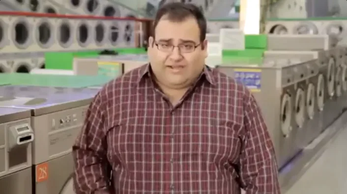 a man in a red plaid shirt and glasses stands in a laundromat surrounded by industrial washing machines, looking slightly…