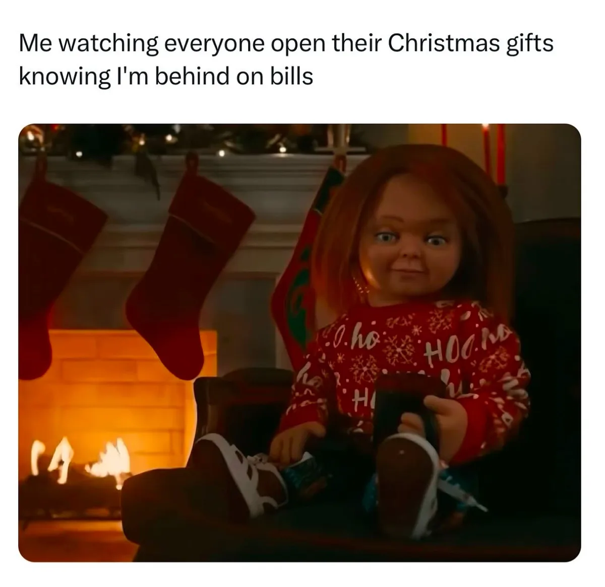 A doll with red hair and a red holiday sweater sits in a chair in front of a lit fireplace, with Christmas stockings hanging…