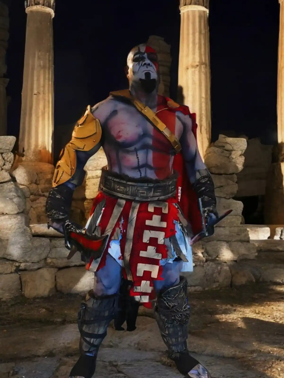 A person in a detailed Kratos cosplay from God of War stands in front of ancient stone pillars at night, holding two blades…