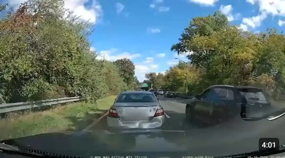 A dashcam view from a car driving on a tree-lined road with moderate traffic under a blue sky. The car directly ahead is a…