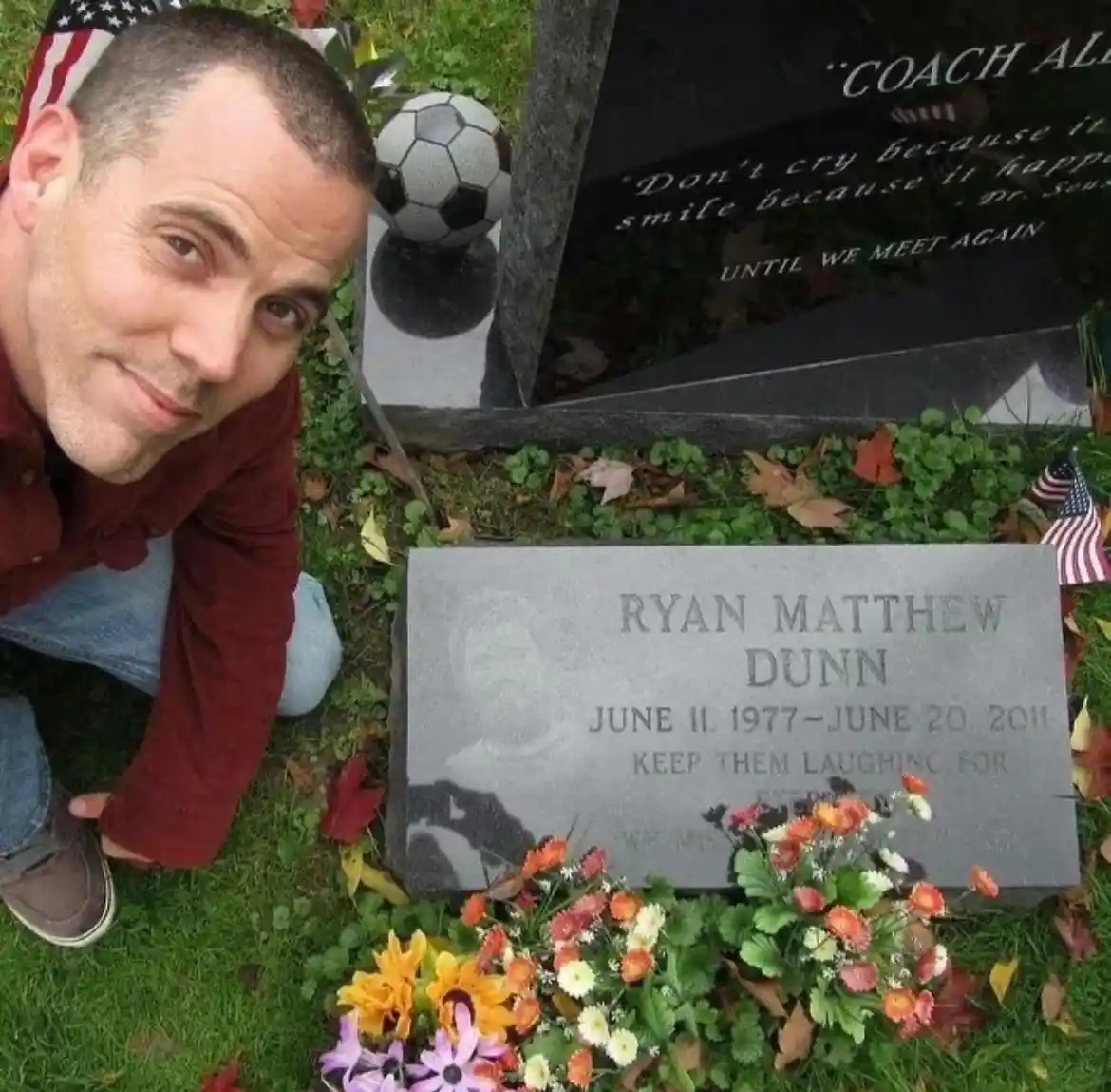 A man in a red shirt and jeans kneels beside a gravestone for Ryan Matthew Dunn, who died in 2011. The grave has a soccer…