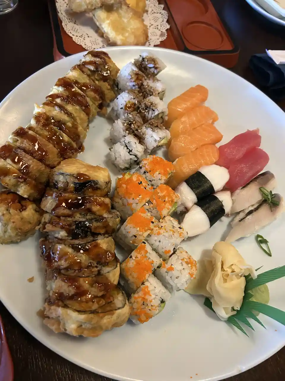 A large white plate is filled with various types of sushi including tempura rolls drizzled with sauce, salmon nigiri, tuna…
