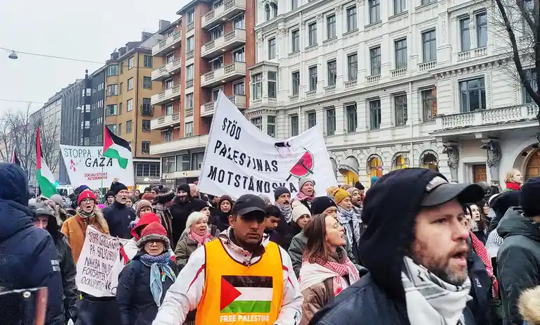 A crowd of protesters marches in a city street, holding signs and Palestinian flags. A large white banner reads 'STÖD…