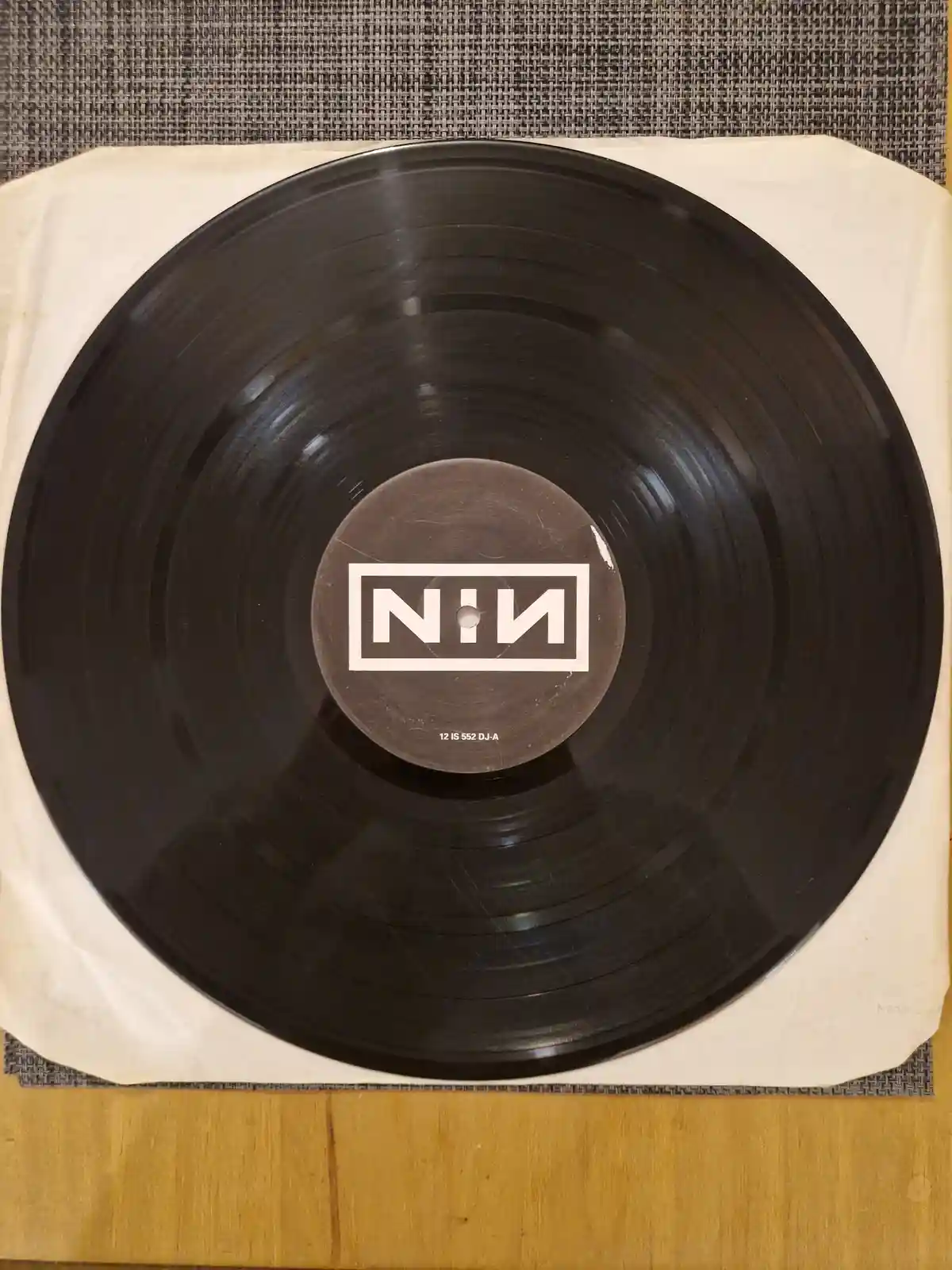 A top-down photo of a black vinyl record resting on a beige paper sleeve, placed on a wooden surface with a woven gray…