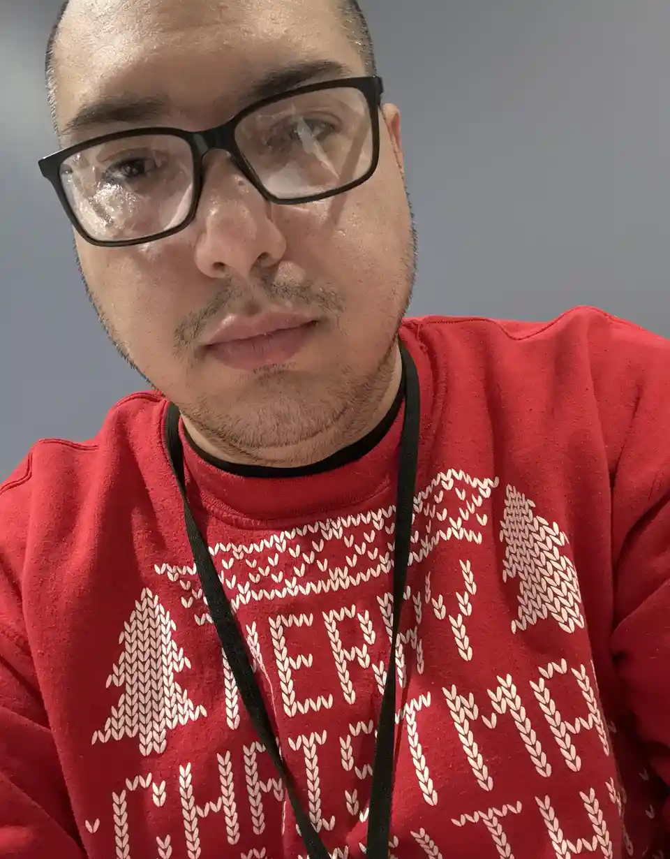 A close-up selfie of a man with short hair and black-rimmed glasses, wearing a red holiday sweater with white knit patterns…