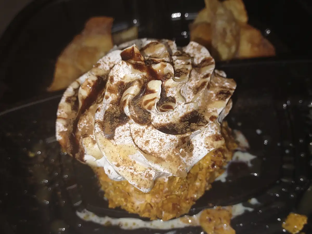 A close-up photo shows a dessert, likely a cupcake, topped with whipped cream, caramel drizzle, and cinnamon powder, served…
