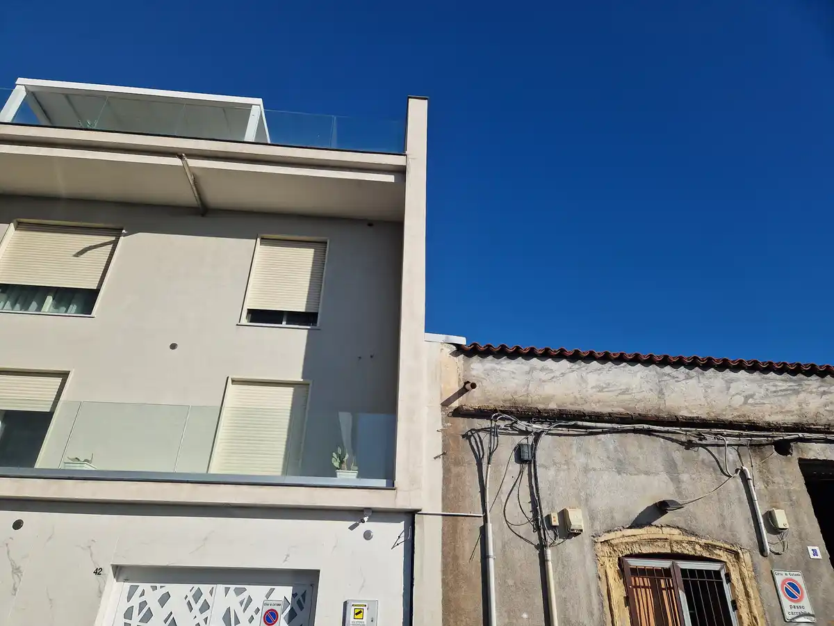A modern, clean white building with glass balconies and closed blinds stands next to a weathered, older building with a…