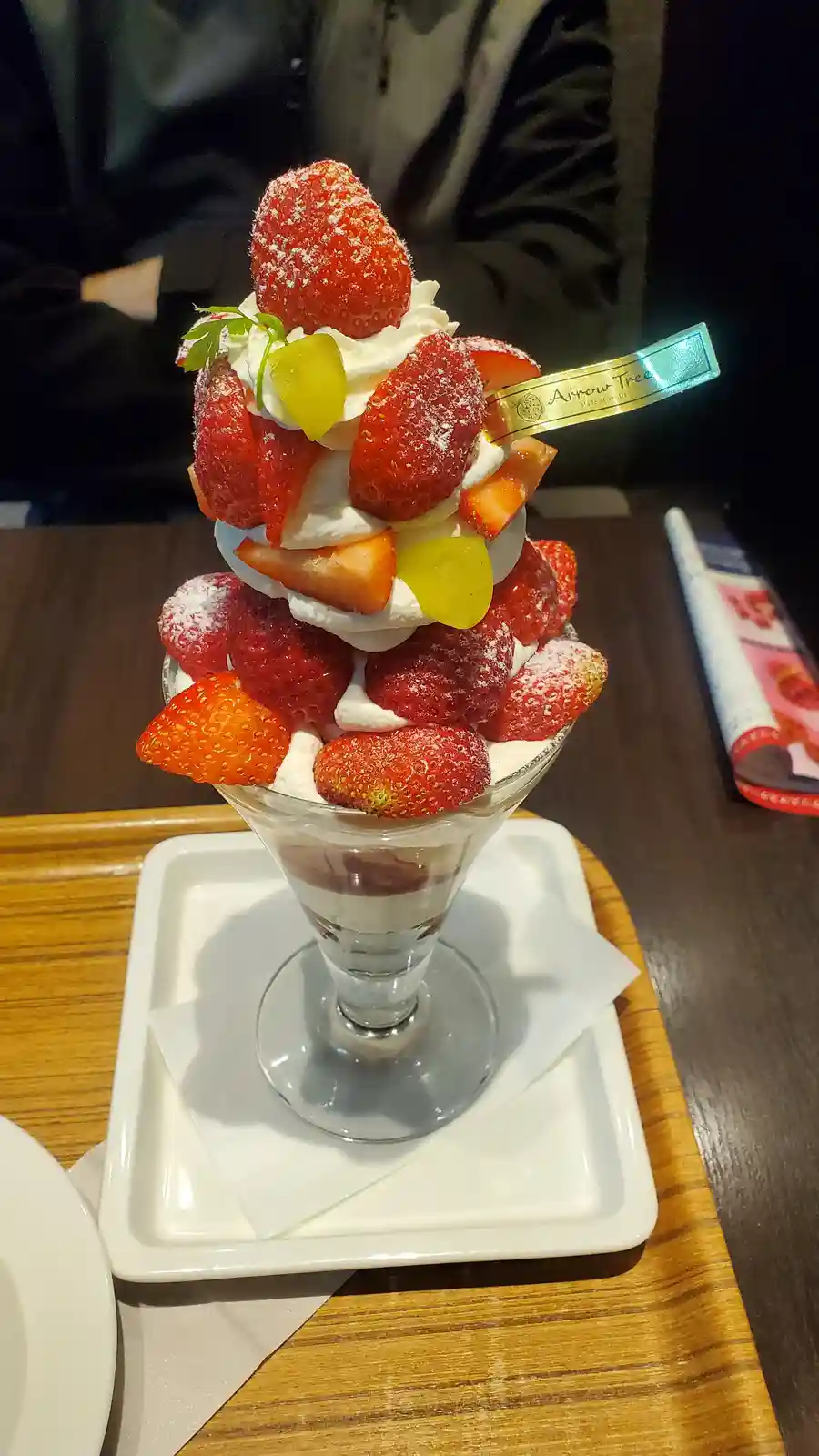 A tall, layered dessert in a glass cup is stacked high with fresh strawberries, whipped cream, and yellow candied fruit…