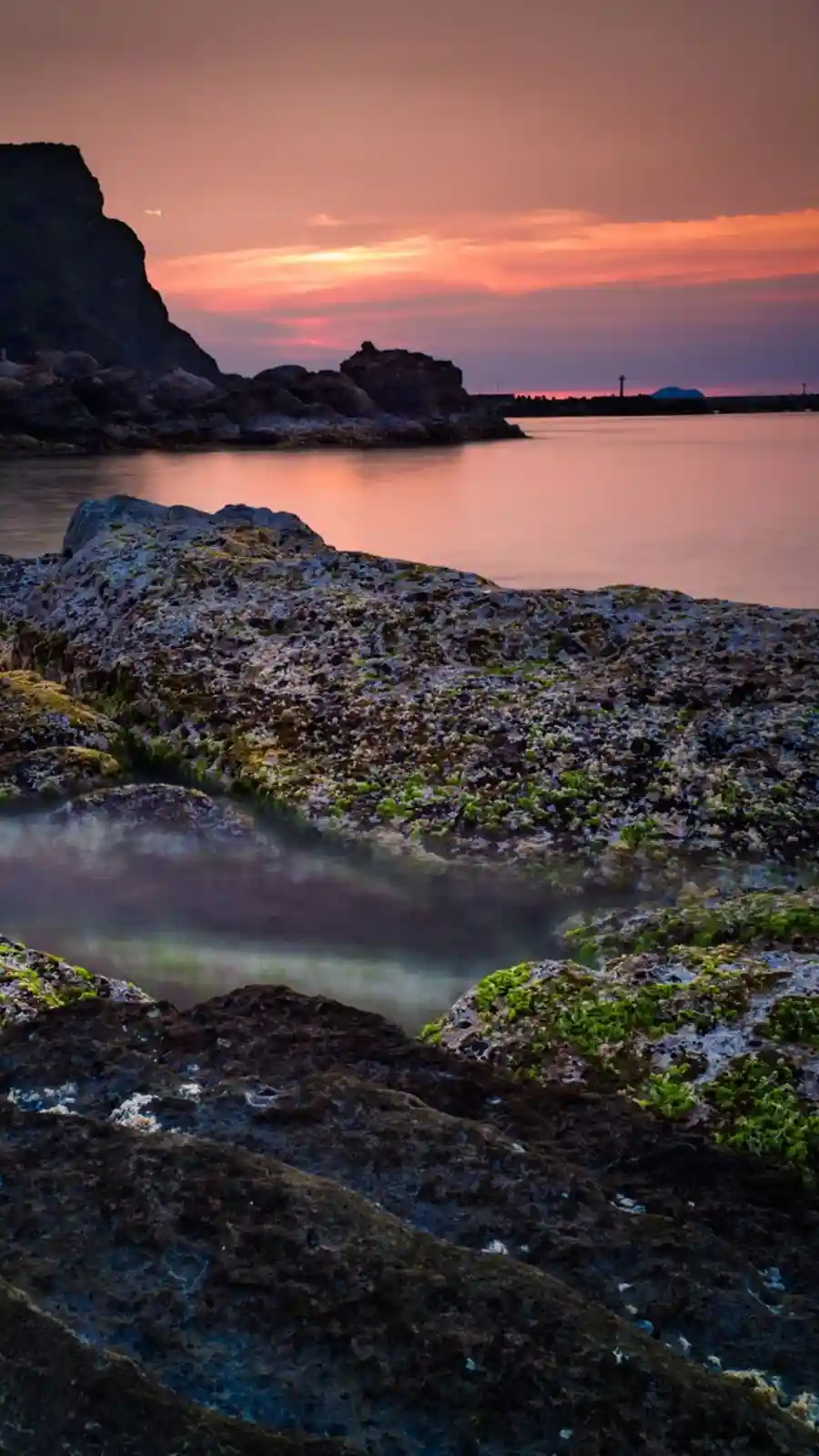 A serene, long-exposure photograph of a rocky shoreline at sunset, with calm water reflecting orange and pink hues in the…